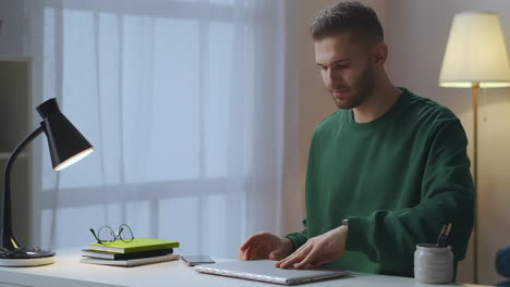young-handsome-man-dressed-green-sweatshirt-is-typing-on-laptop-internet-user-is-working-alone-in-apartment-connecting-to-net-and-turning-on-table-lamp-medium-shot